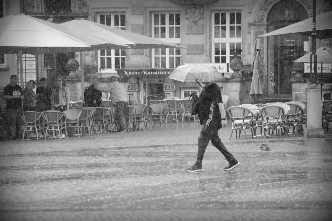 A monochrome photograph oif a cobblestoned market place, surrounded by the outdoor tables and sunshades of restaurants, with a single person hurrxying across the centre under an umbrella in the pouring rain. Copyright Urban Camera.