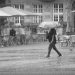 A monochrome photograph oif a cobblestoned market place, surrounded by the outdoor tables and sunshades of restaurants, with a single person hurrxying across the centre under an umbrella in the pouring rain. Copyright Urban Camera.
